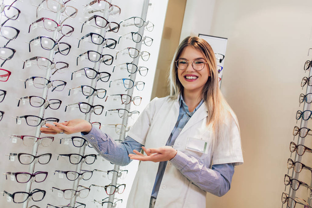 Aantrekkelijke jonge vrouwelijke arts in oogheelkunde kliniek. Oogarts dokter staat in de buurt van planken met verschillende brillen. - Foto, afbeelding