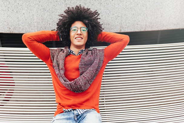 A handsome young Egyptian curly guy sitting on a bench dressed in a bright orange sweater and jeans. Happy people. The student walks in the break between the lessons. - Photo, image