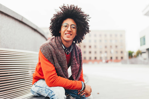 A handsome young Egyptian curly guy sitting on a bench dressed in a bright orange sweater and jeans. Happy people. The student walks in the break between the lessons. - Foto, afbeelding