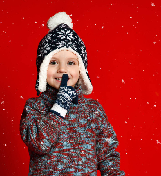 child boy in knitted hat and sweater and mittens making silence gesture  over colorful red  background. - Foto, Imagem