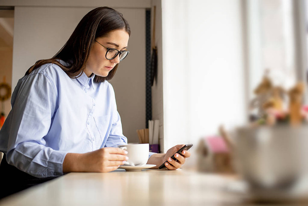 Vrouw met donker haar, bril, blauwe shirt zitten in een cafe, koffie drinken en kijken naar de telefoon - Foto, afbeelding