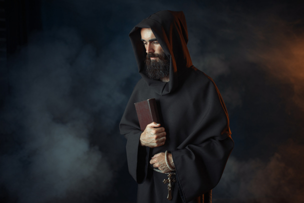 Medieval monk in robe holds spellbook in hands, black background, secret ritual. Mysterious friar in dark cape. Mystery and spirituality - Фото, изображение