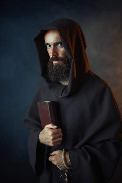 Medieval monk in robe holds spellbook in hands, black background, secret ritual. Mysterious friar in dark cape. Mystery and spirituality - Foto, afbeelding