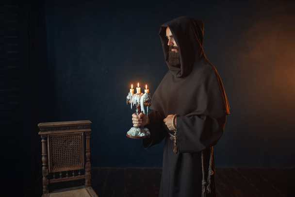 Medieval monk in robe holds a candlestick in hands, black background, secret ritual. Mysterious friar with candles. Mystery and spirituality - Photo, Image