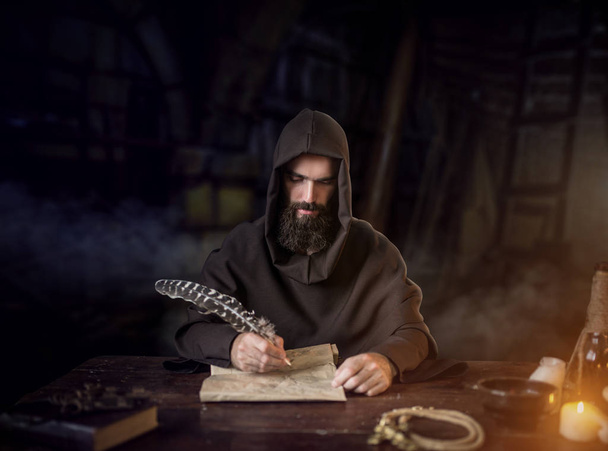 Mittelalterlicher Mönch in Gewand und Kapuze schreibt mit Gänsefeder, schwarzem Hintergrund, geheimen Ritual. geheimnisvoller Mönch in dunklem Umhang. Geheimnis und Spiritualität - Foto, Bild