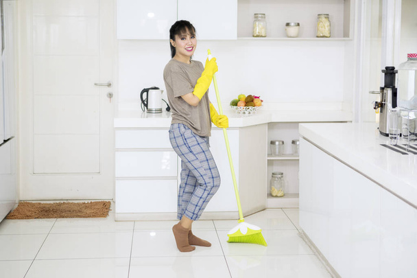 Picture of young housekeeper cleaning floors with a broom while standing in the kitchen - Zdjęcie, obraz