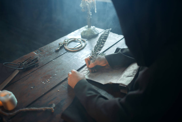 Monje medieval sentado en la mesa y escribe con una pluma de ganso, vista superior, escritura secreta. Misterioso fraile de capa oscura. Misterio y espiritualidad
 - Foto, imagen