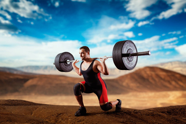 Muscular weightlifter doing squats with a barbell, deadlift, sandy desert valley on background. Weightlifting workout outdoor, bodybuilding training - Photo, Image