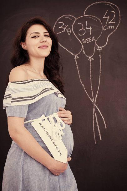 Portrait of happy pregnant woman with hands on belly in front of black chalkboard - Photo, image