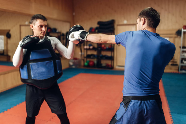 Mannelijke kickbokser in handschoenen hand punch met een persoonlijke trainer in pads, sporten in de fitnessruimte beoefenen. Boxer op opleiding, kickboksen praktijk - Foto, afbeelding