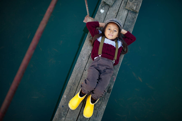 Dziecko dziewczynka w WPR i żółte kalosze leży na drewniany most i uśmiechów na tle rzeki. Widok z góry. - Zdjęcie, obraz