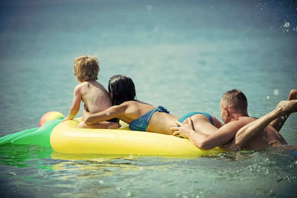 Family vacation concept. Father swim near, pushes mattress. Happy young family spend time together and having fun. Cute child boy sits on air mattress pineapple shaped with mother, in ocean, sea. - Φωτογραφία, εικόνα