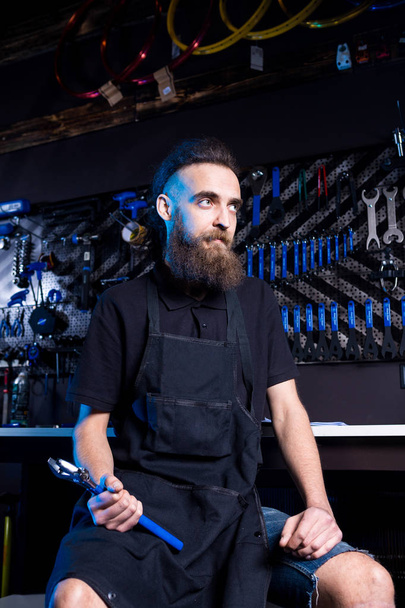 Portrait of small business owner of young man with beard. Guy bicycle mechanic workshop worker sitting with tool in his hand in a working black clothes in an apron in the background of a bicycle shop. - Fotoğraf, Görsel