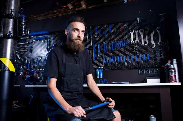 Retrato do pequeno empresário do jovem com barba. Guy trabalhador oficina mecânica de bicicleta sentado com ferramenta em sua mão em uma roupa preta trabalhando em um avental no fundo de uma loja de bicicletas
. - Foto, Imagem