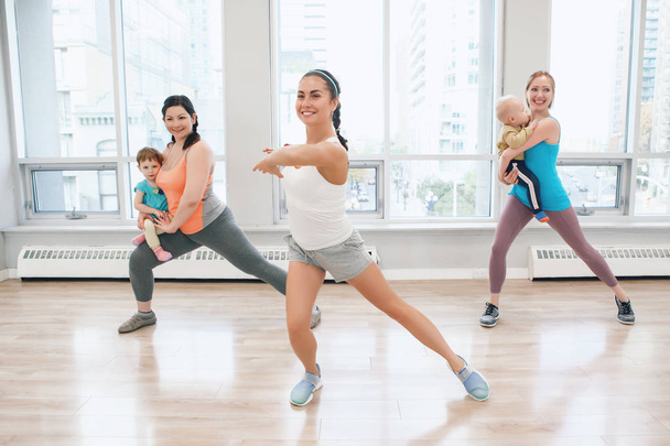 Group of three young women with children doing workout in gym class with instructor to loose baby weight. Child-friendly fitness for mothers with kids toddlers. Lifestyle concept of parent activity - Photo, Image
