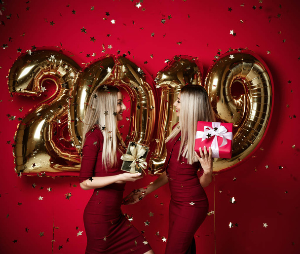 Two women celebrating at new year party happy laughing girls in casual dresses throw gold stars confetti with 2019 balloons - Foto, imagen