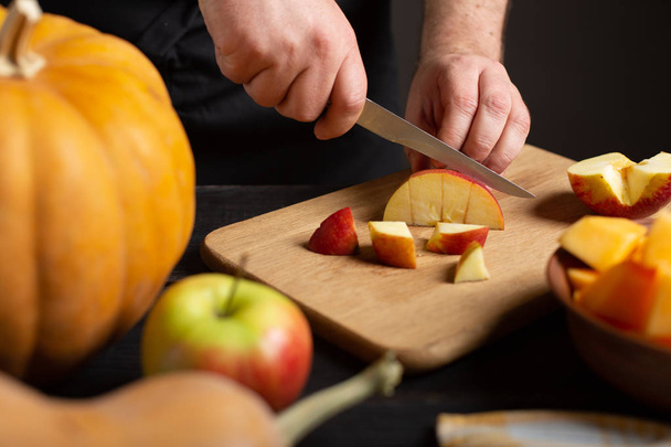 Ο μάγειρας κόβει το μήλο σε κομμάτια για το ψήσιμο. Σε ένα ξύλινο τραπέζι μαύρο ψέμα κολοκύθες διαφόρων μεγεθών και σχημάτων, ώριμα μήλα, μια πετσέτα για τα πιάτα και ένα μπολ φέτες κολοκύθας. - Φωτογραφία, εικόνα