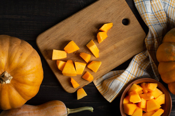 On a wooden black table lie pumpkins of various sizes and shapes, a dishcloth and a bowl of sliced pumpkin. In the center of the frame is a cutting board with sliced pumpkin. Top view. - 写真・画像