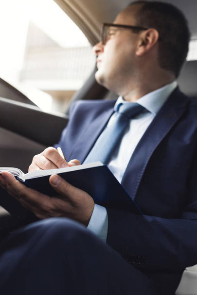 Man in a business suit write on notebook with laptop in the salon of an expensive car with leather interior - Fotó, kép