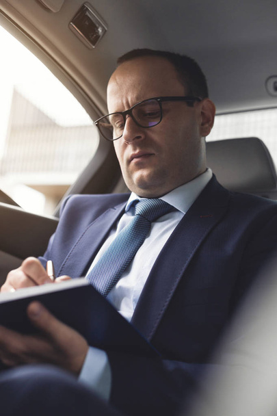 Man in a business suit write on notebook with laptop in the salon of an expensive car with leather interior - Photo, image