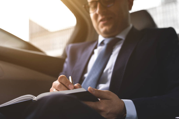 Man in a business suit write on notebook with laptop in the salon of an expensive car with leather interior - Photo, image