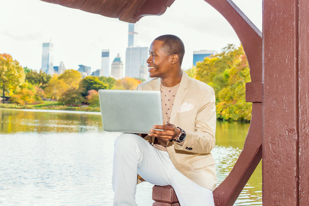 Geschäftsmann arbeitet. In cremefarbenem Blazer, kragenlosem Pullover, weißer Hose, mit Armbanduhr sitzt ein junger Schwarzer am See in der Großstadt, lächelt, denkt, arbeitet am Laptop. - Foto, Bild