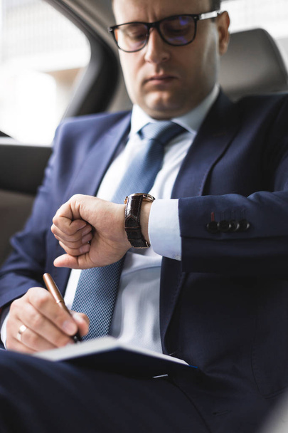 Man in a business suit write on notebook with laptop in the salon of an expensive car with leather interior - Photo, Image