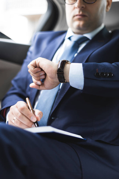 Man in a business suit write on notebook with laptop in the salon of an expensive car with leather interior - Фото, изображение