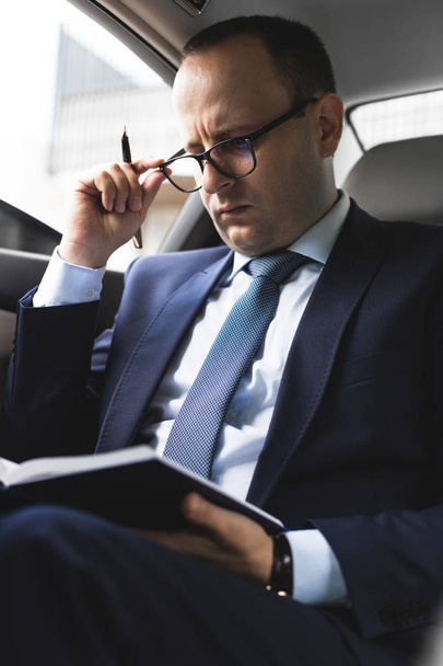 Man in a business suit write on notebook with laptop in the salon of an expensive car with leather interior - Фото, зображення