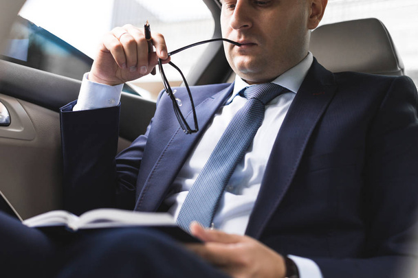 Man in a business suit write on notebook with laptop in the salon of an expensive car with leather interior - Fotografie, Obrázek