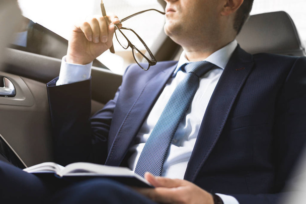 Man in a business suit write on notebook with laptop in the salon of an expensive car with leather interior - Фото, зображення
