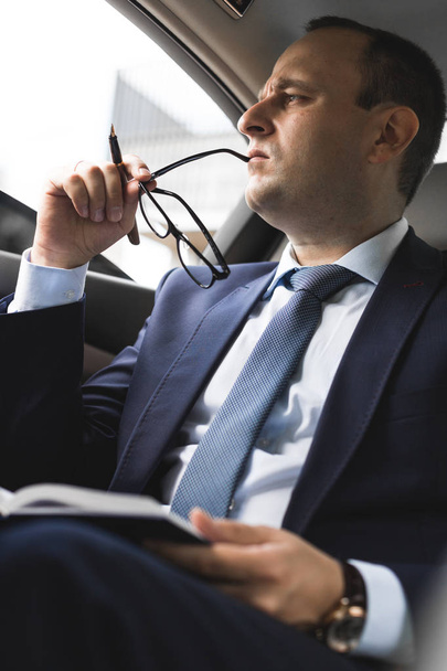 Man in a business suit write on notebook with laptop in the salon of an expensive car with leather interior - Foto, Imagem
