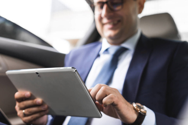 Young businessman using tablet pc while sitting on back seat of a car. Caucasian male business executive by a taxi and looking at digital tablet. - Foto, Imagem