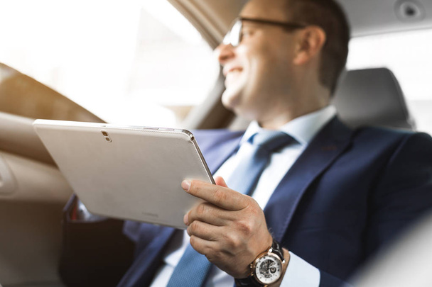 Young businessman using tablet pc while sitting on back seat of a car. Caucasian male business executive by a taxi and looking at digital tablet. - Foto, imagen