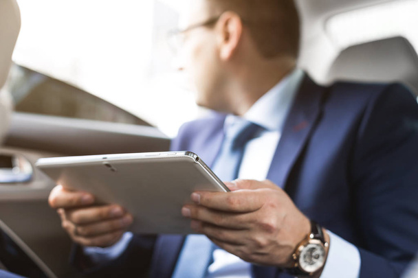 Young businessman using tablet pc while sitting on back seat of a car. Caucasian male business executive by a taxi and looking at digital tablet. - Valokuva, kuva