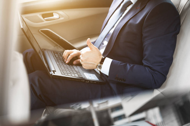 ein junger Geschäftsmann im Anzug sitzt mit Laptop und Notizbuch auf dem Rücksitz eines Geschäftsautos, Geschäftsverhandlungen - Foto, Bild