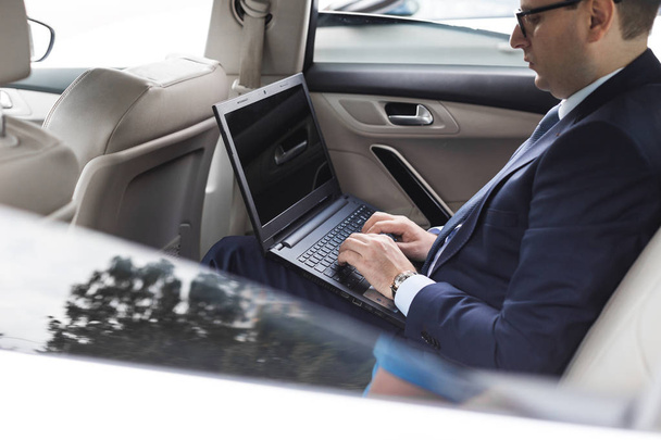 a young business man in a suit is sitting in the back seat of a business car with a laptop and a notebook, business negotiations - Foto, imagen