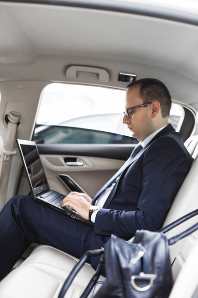 a young business man in a suit is sitting in the back seat of a business car with a laptop and a notebook, business negotiations - Valokuva, kuva