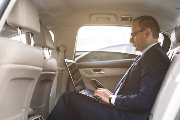a young business man in a suit is sitting in the back seat of a business car with a laptop and a notebook, business negotiations - Fotoğraf, Görsel