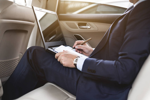 a young business man in a suit is sitting in the back seat of a business car with a laptop and a notebook, business negotiations - Φωτογραφία, εικόνα