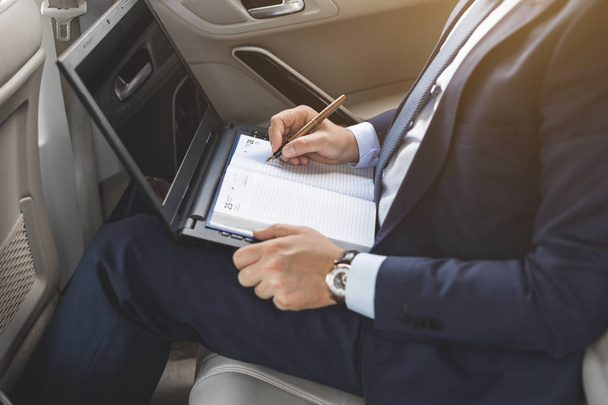 a young business man in a suit is sitting in the back seat of a business car with a laptop and a notebook, business negotiations - Zdjęcie, obraz