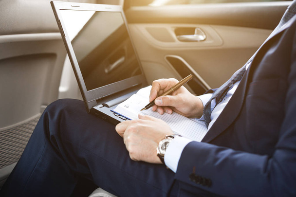 a young business man in a suit is sitting in the back seat of a business car with a laptop and a notebook, business negotiations - Foto, Bild