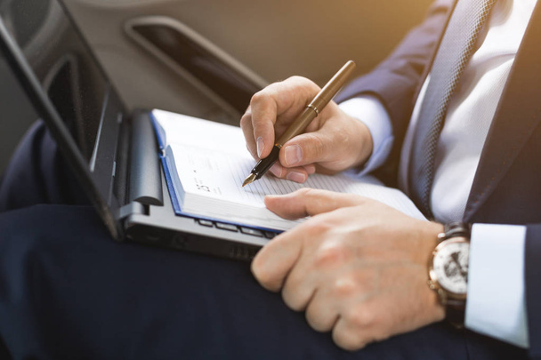um jovem homem de negócios em um terno está sentado no banco de trás de um carro de negócios com um laptop e um notebook, negociações de negócios
 - Foto, Imagem