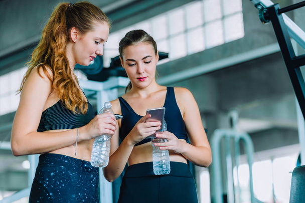 Fit beautiful young woman athletes in fitness clothes relaxing after workout in gym. Couple women standing holding water in bottle and using fitness app smartphone. Healthy, Sport, Lifestyle concept. - Photo, Image