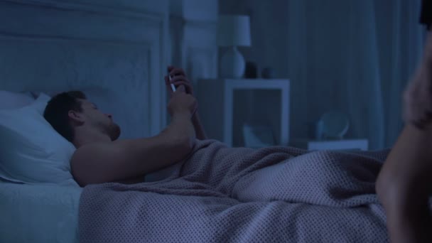 Man lying in bed chatting on smartphone, girlfriend coming in seductive lingerie - Video, Çekim