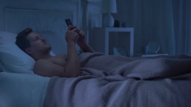 Man lying in bed holding phone, turning away from woman coming in lingerie - Πλάνα, βίντεο