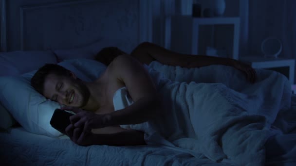 Male in bed using dating site while wife sleeping, lady looking at smartphone - Πλάνα, βίντεο