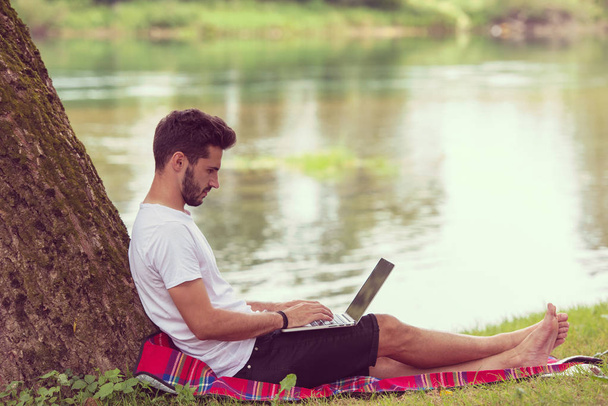 Um jovem freelancer usando um computador portátil enquanto trabalhava em bela natureza sob a árvore na margem do rio
 - Foto, Imagem