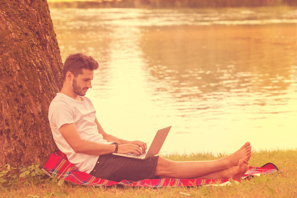 A young freelancer using a laptop computer while working in beautiful nature under the tree on the bank of the river - Foto, Imagen