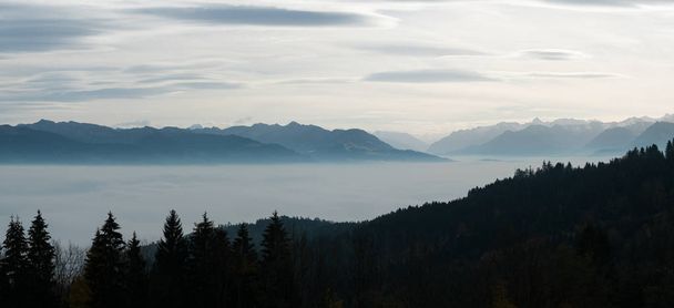 катящиеся горные хребты в легком туманном небе с осенним цветным лесом на переднем плане и многими заснеженными горами позади
 - Фото, изображение